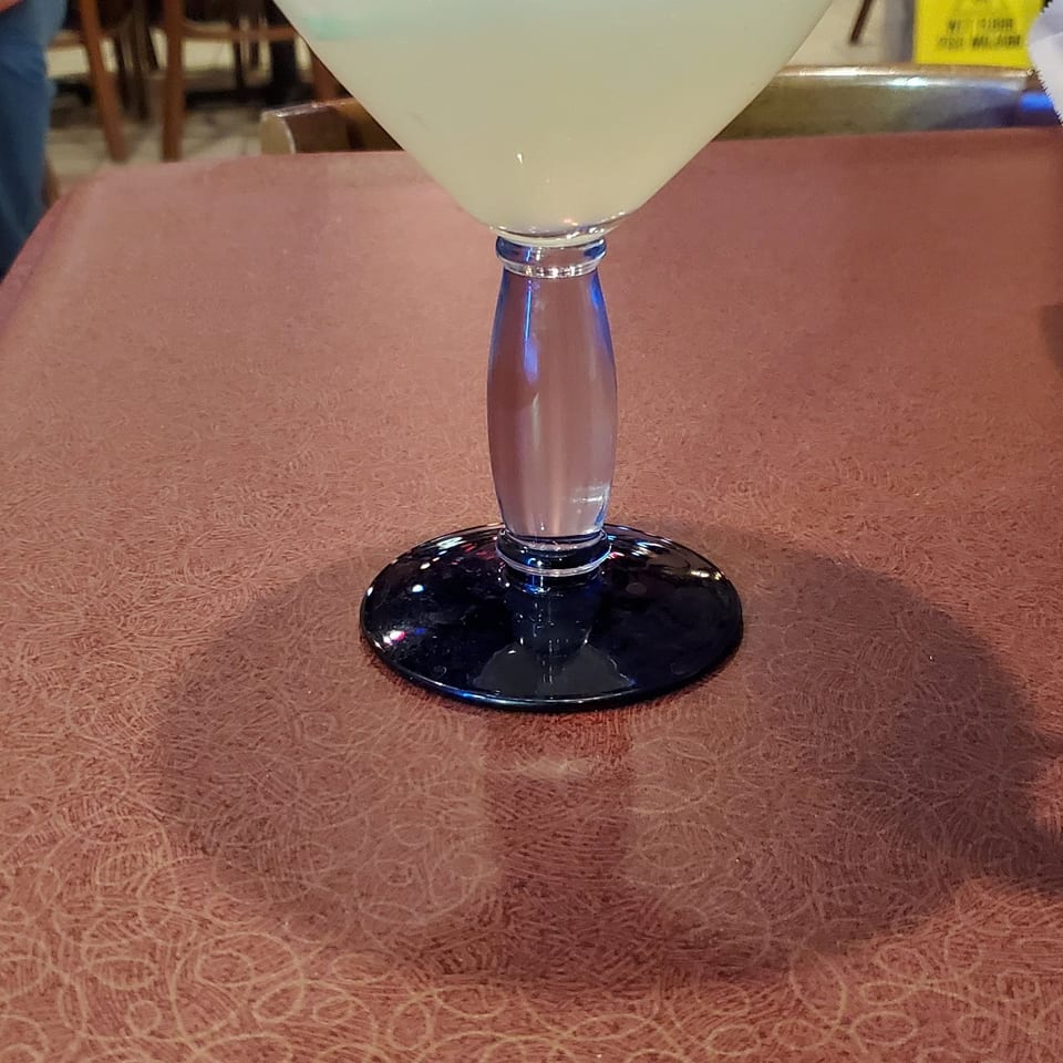 Large pale margarita in a blue-rimmed glass with a lime wedge, set on a restaurant table with diners in the background.