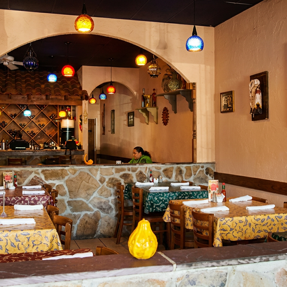 Warmly lit restaurant dining room with colorful pendant lamps, patterned tablecloths, stone accents, and a bar stocked with bottles.