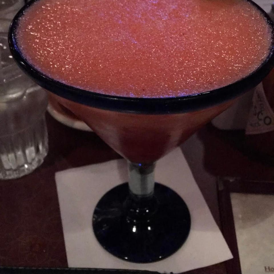 Pink frozen cocktail in a margarita glass with a salted rim, lime wedge, and red straw on a restaurant table.