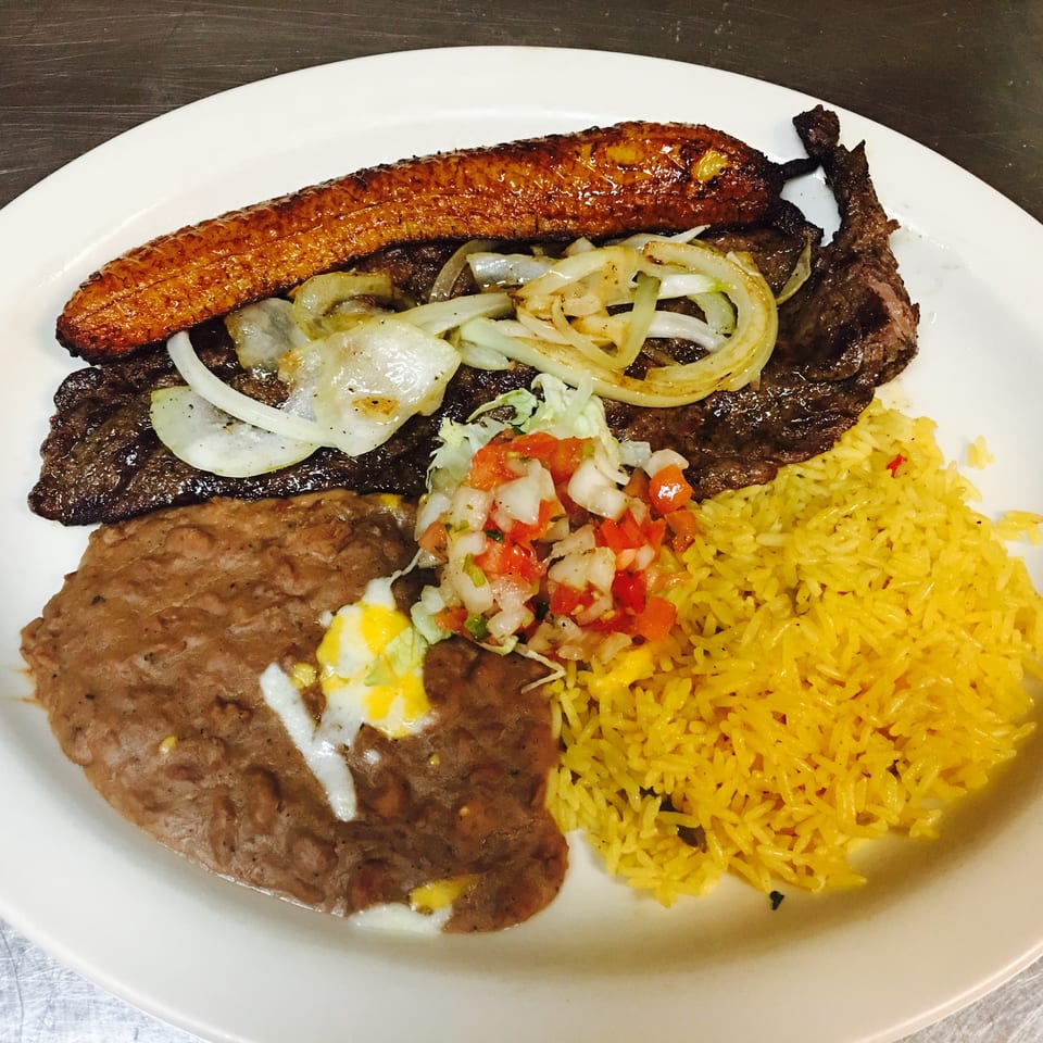 Plate of grilled steak, fried plantain, yellow rice, beans, sautéed onions, and fresh tomato salsa.